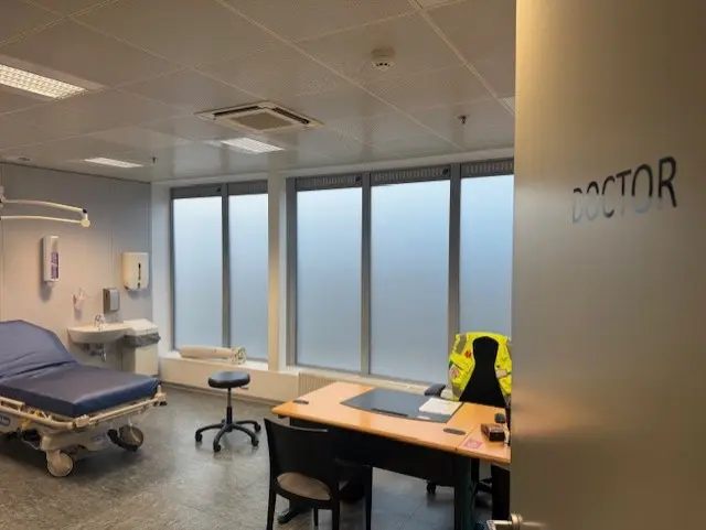 A workplace medical room signed DOCTOR, with an examination trolley, washbasin, wall-mounted dispensers and a desk