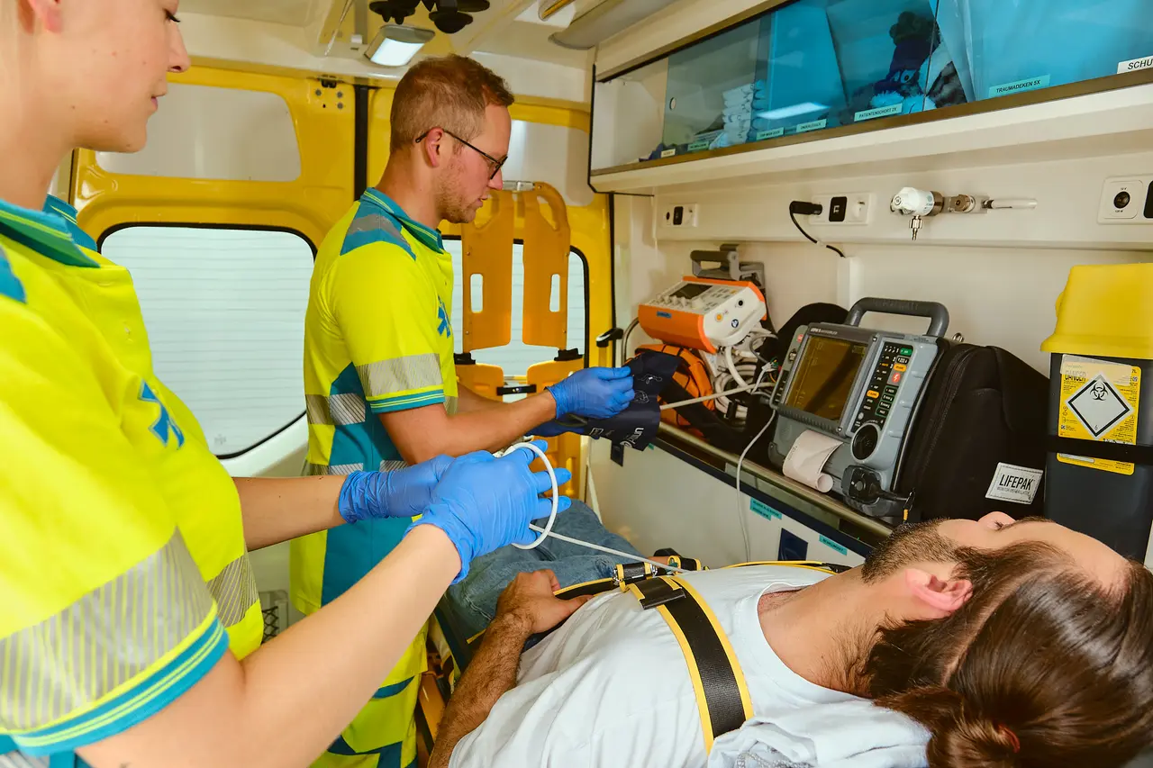 Two paramedics in high-visibility uniforms treating a patient secured to a stretcher inside an ambulance patient compartment, with a defibrillator monitor and stocked cabinets alongside